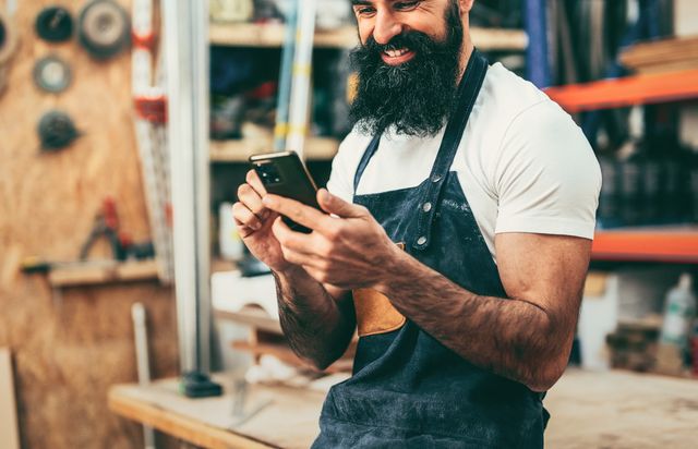 A bald, bearded woodworker in a navy apron in his workshop, grinning at his phone