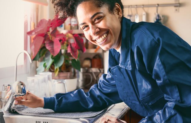 A plumber in blue overalls at a customer’s kitchen sink, laughing as she checks the tap