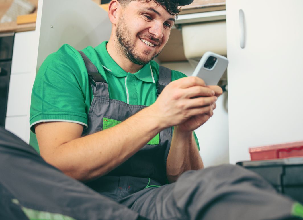 A plumber sitting on a customer’s kitchen floor mid-job, grinning at his phone